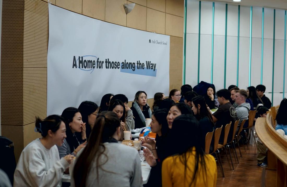 Members of ARK Church Seoul eating together at long tables beneath a banner reading A Home for those along the Way