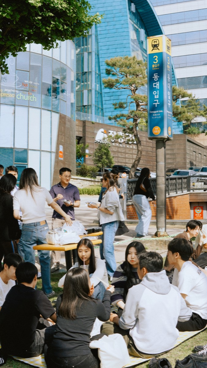 Members of ARK Church Seoul sitting on the grass beside the Dongguk University Station Exit 3 sign, with the church building behind