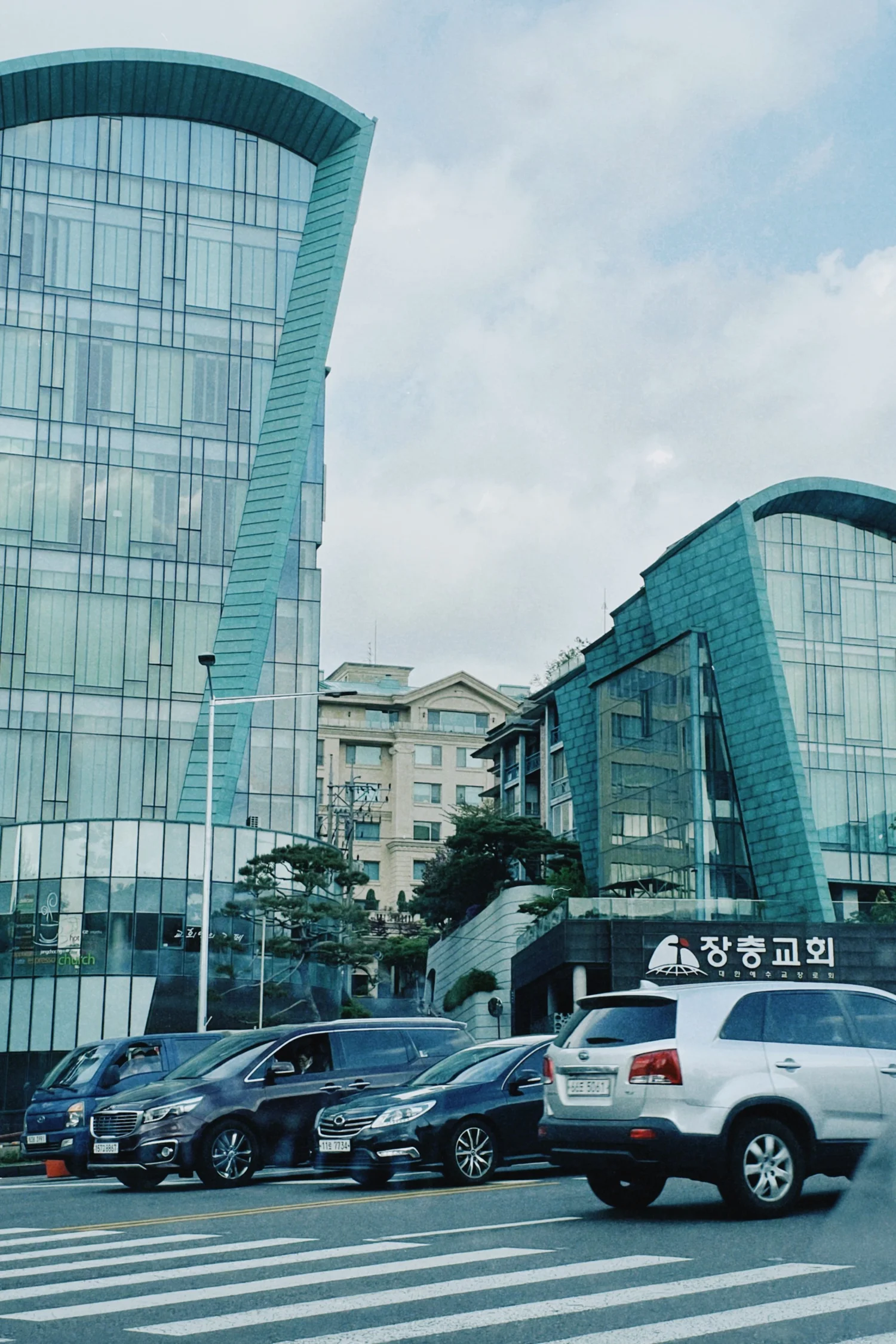 IN SEOUL SOUTH KOREA ENGLISH CHURCH GREAT LOCATION Modern glass buildings with curved architectural design, parked cars, and signage in Korean, with a crosswalk in the foreground.