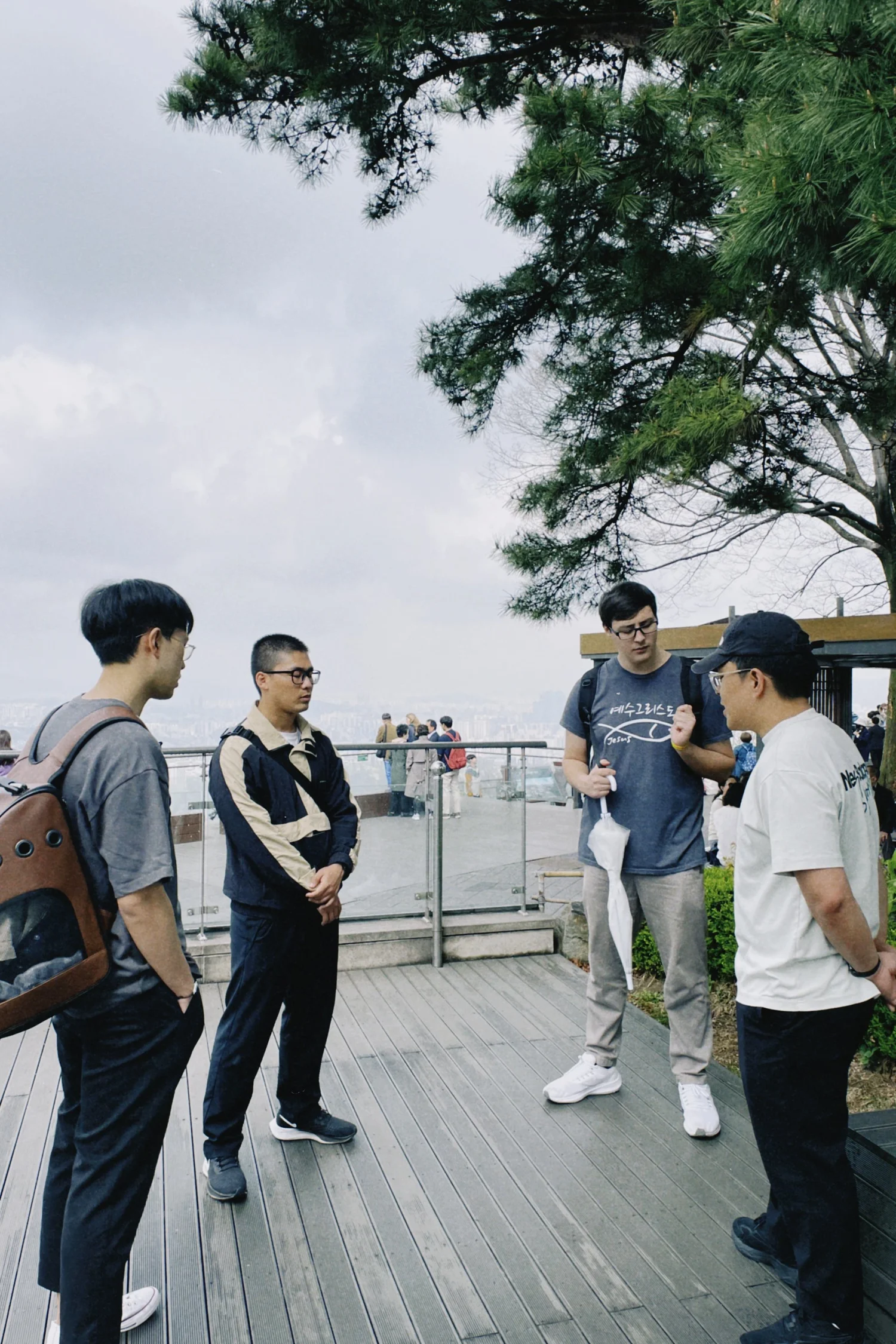 A group of five young men standing outdoors on a wooden platform, engaged in conversation with one another. There is a tree with green foliage and a cloudy sky in the background.IN SEOUL SOUTH KOREA ENGLISH CHURCH OUTING