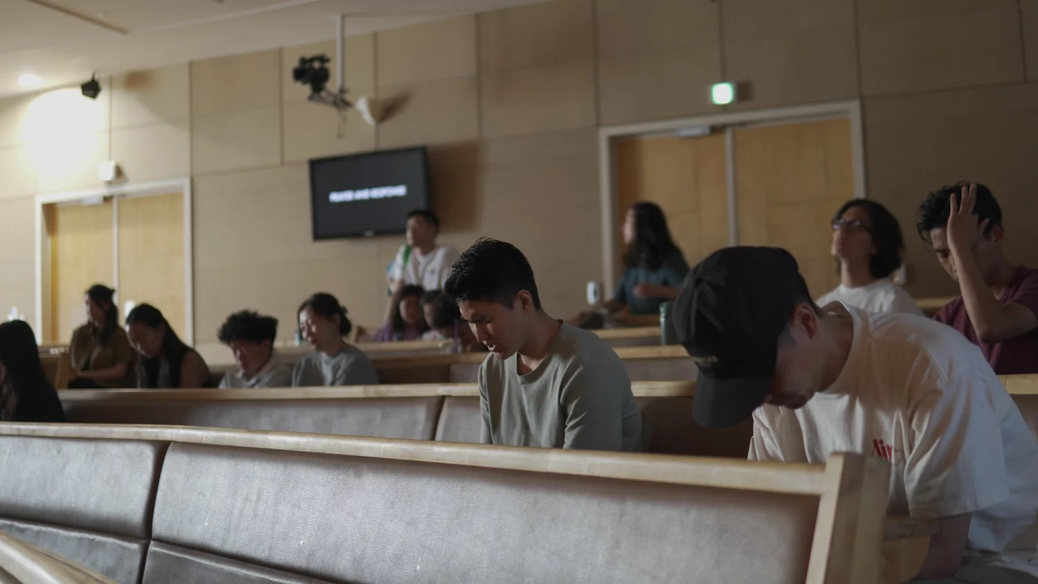 A group of people sitting in a lecture hall or conference room, some with heads bowed or eyes closed, possibly praying or reflecting. Several attendees are visible, with others standing near the back of the room. There is a screen on the wall displaying some text.
