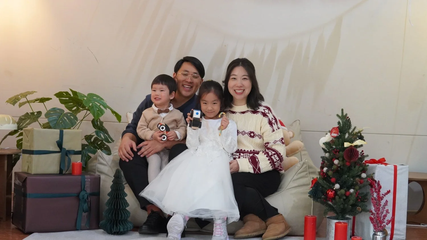 Family of four sitting on a sofa in an English international church in Seoul during Christmas celebration, with decorated Christmas tree and wrapped presents nearby.